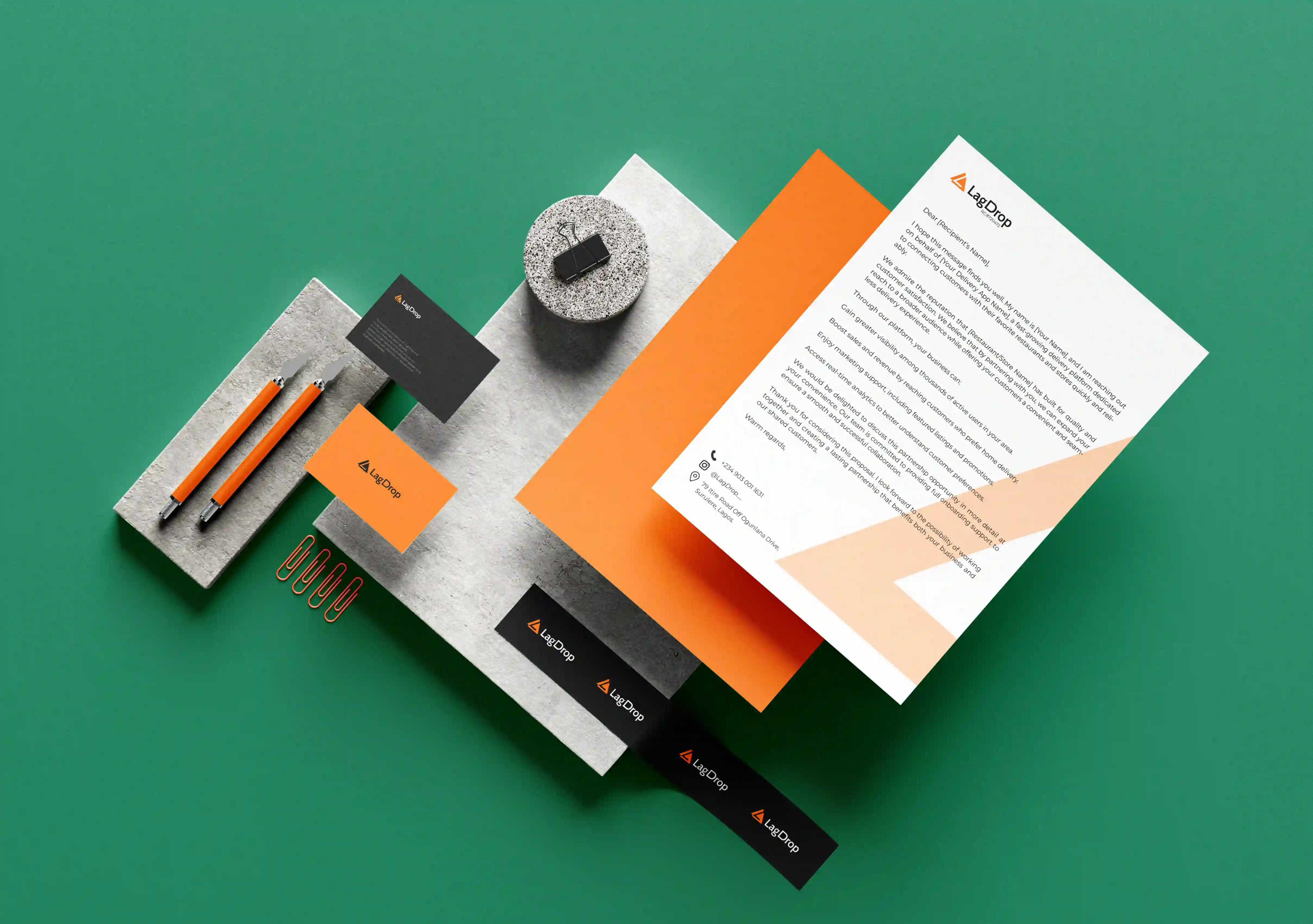 Flat lay of green workspace with orange and black LagDrop branded stationery including pens, business cards, letterhead, clips, and labels.
