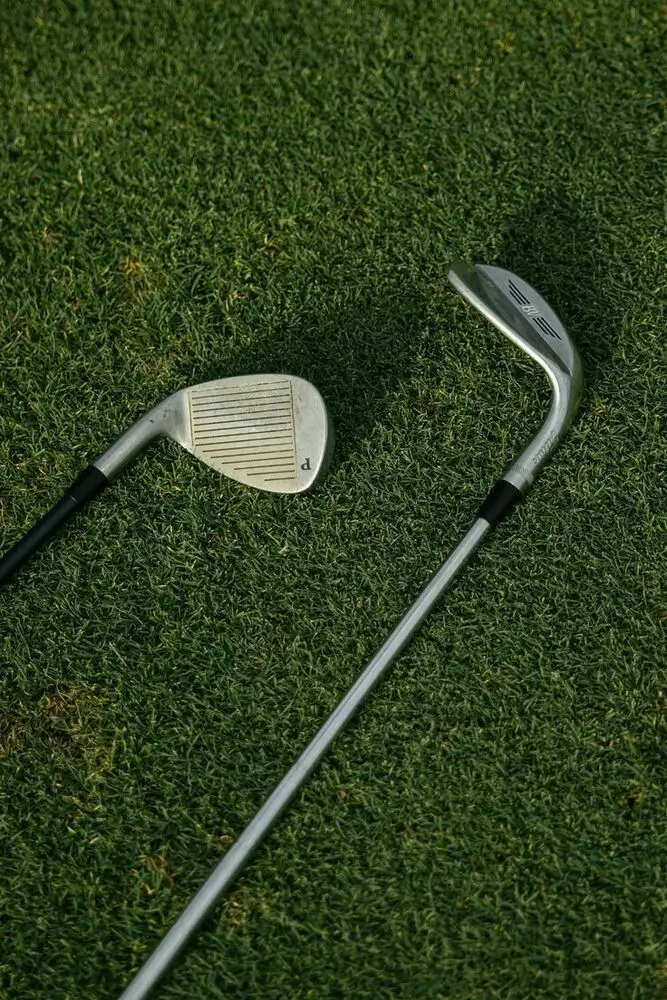 Twee metalen golfclubs liggend op groen gras, één met het gezicht omhoog en één met de steel zichtbaar.