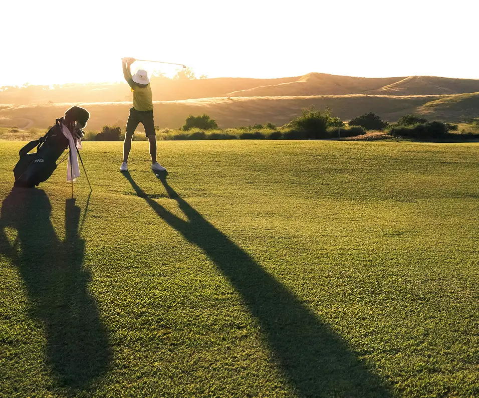 Golfer in voorbereiding op de swing bij zonsondergang op een golfbaan met een golftas naast zich.