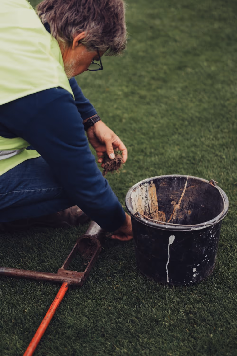 Man aan het knielen op gras met een stuk gras en aarde in zijn hand, naast een zwarte emmer en een tuingereedschap.