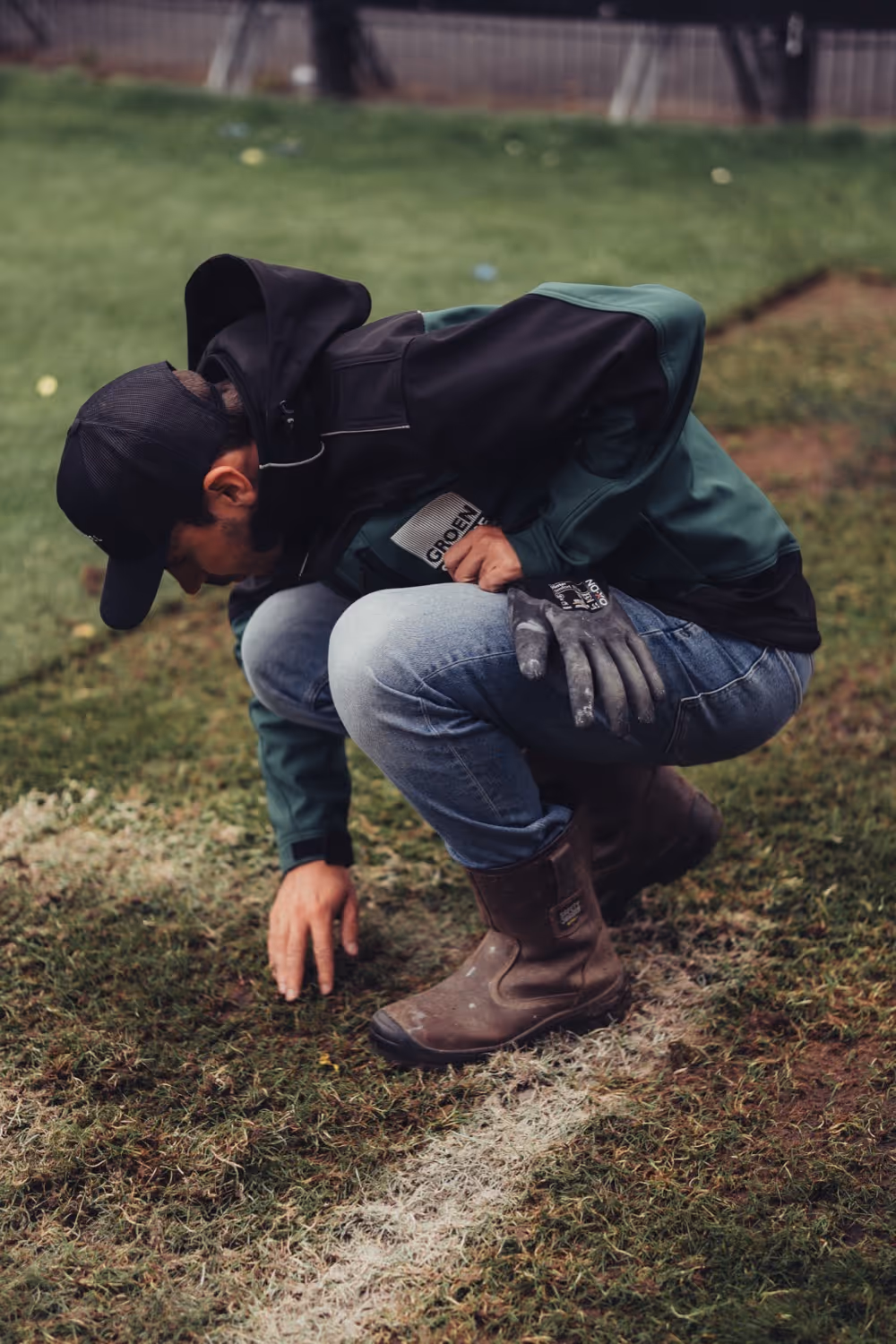 Man in werklaarzen en jas hurkt op het grasveld en onderzoekt de grond langs een witte lijn.