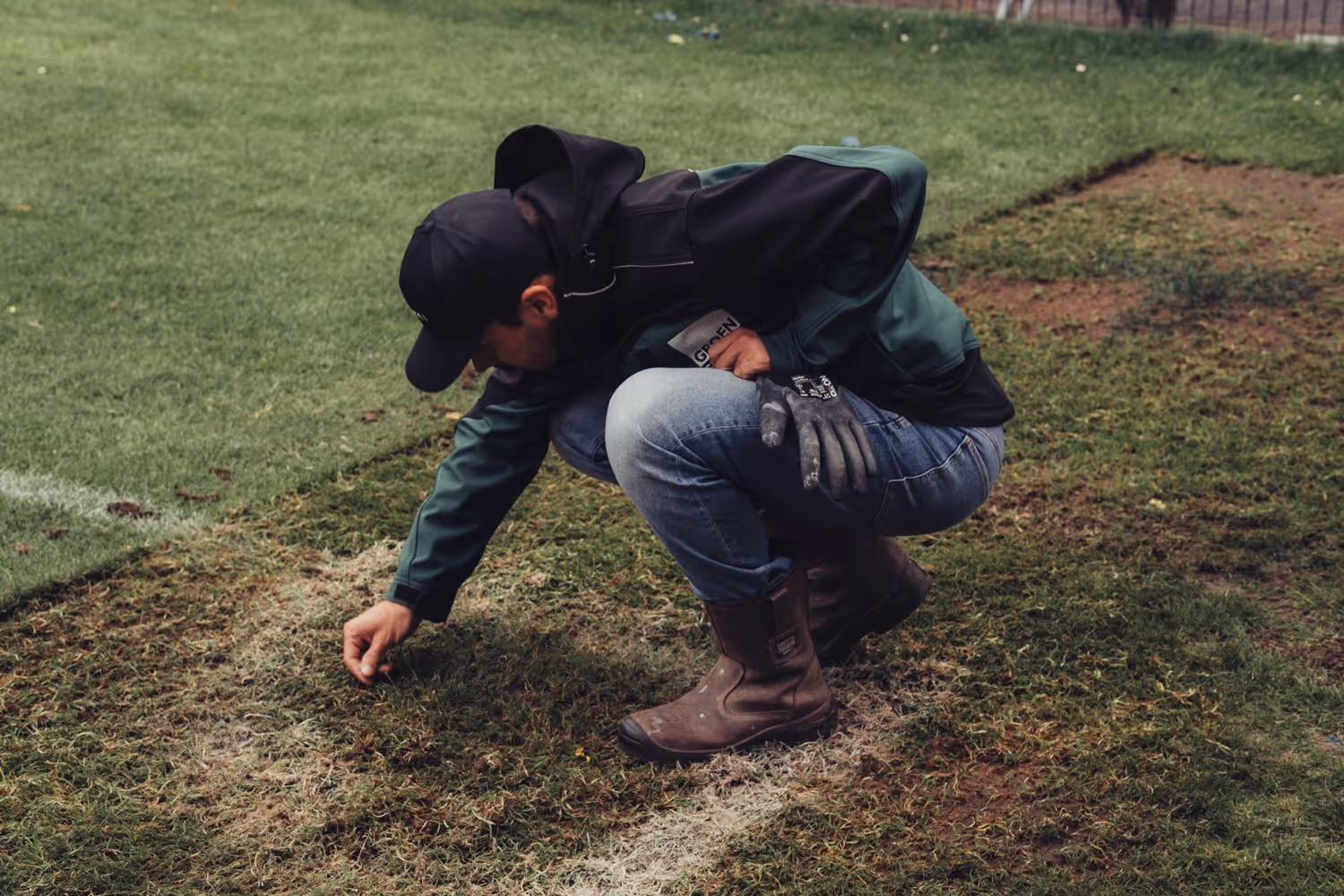 Man in work boots, jeans, and jacket crouching on grass, inspecting or planting near a white line on a field.