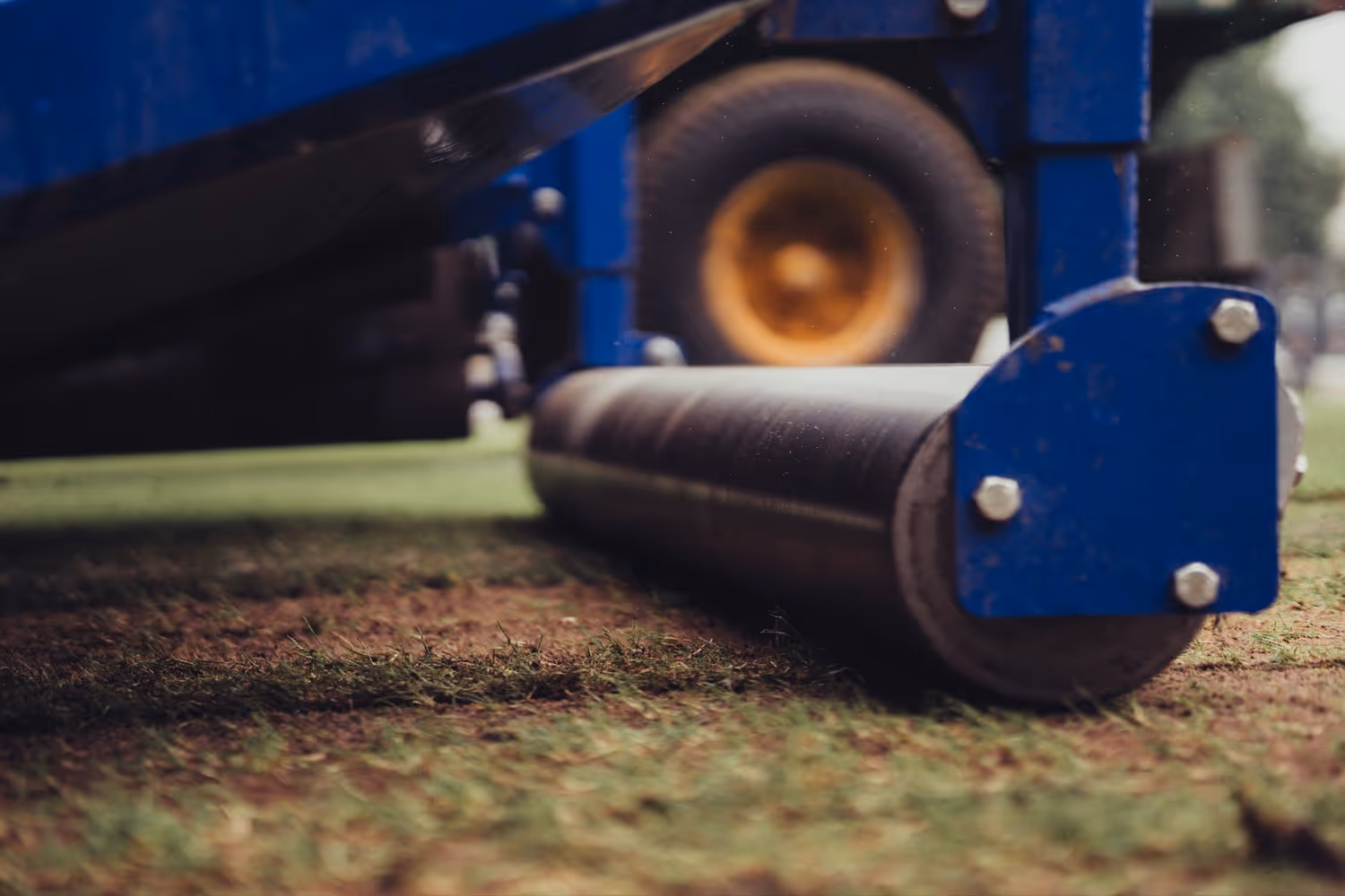 Blauwe machine met rol die gras op een sportveld gladstrijkt.