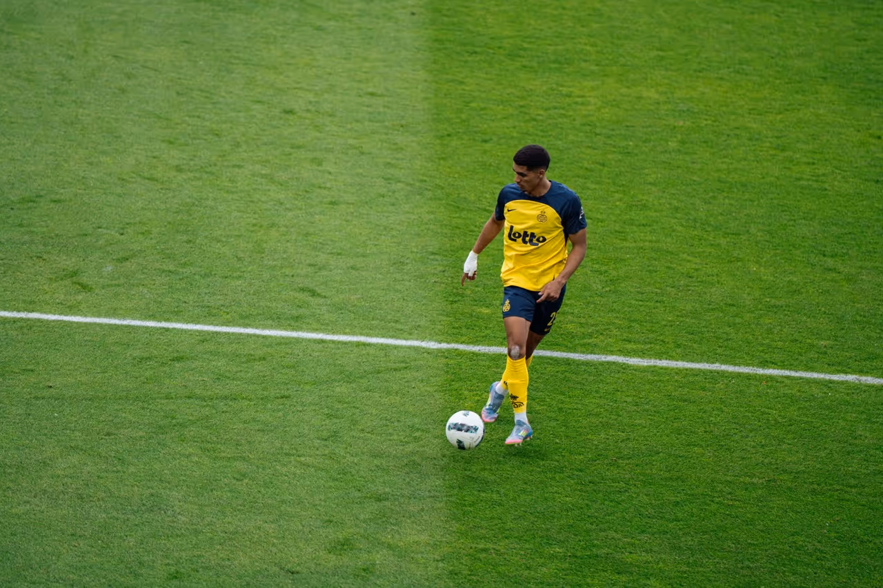 Voetballer in geel-blauw tenue met bal op het grasveld, dicht bij een witte lijn.