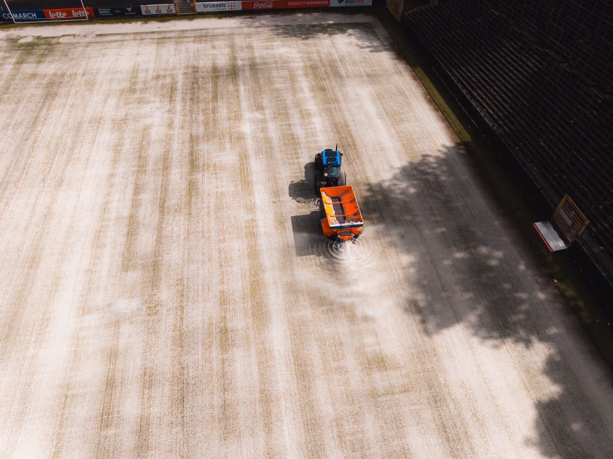 Een kleine tractor met een oranje strooiwagen die zout strooit op een sportveld.