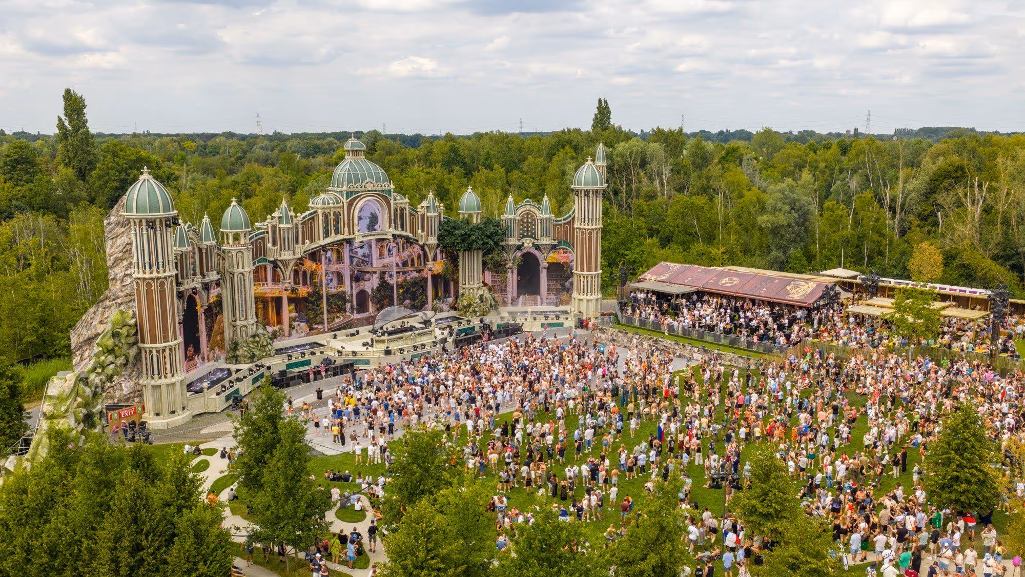 Luchtfoto van een groot buitenfestival met een drukbezochte openluchtpodium omringd door groene bomen onder een bewolkte hemel.