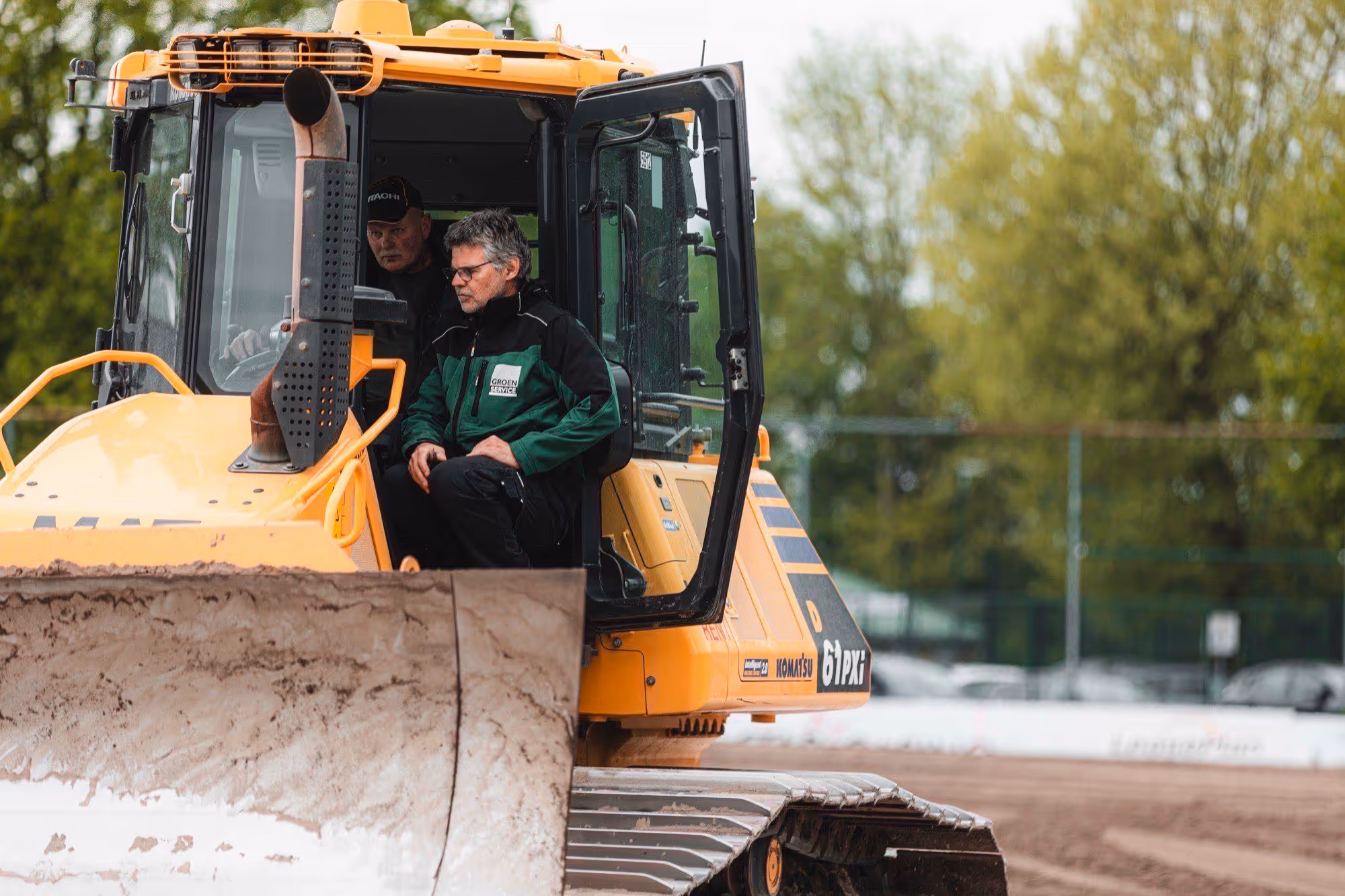 Twee mannen in een gele graafmachine met open deur op een bouwterrein met bomen op de achtergrond.