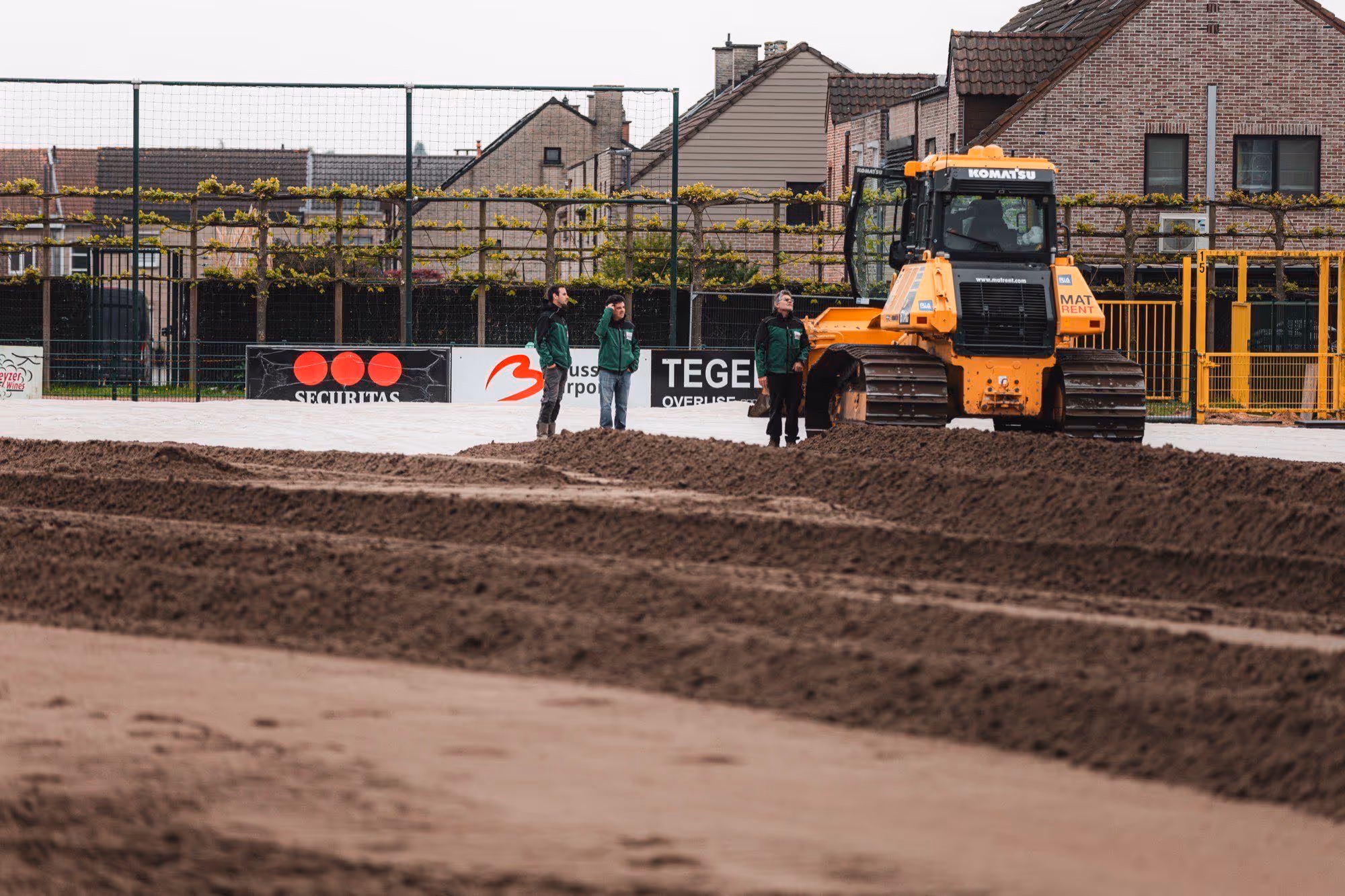 Drie mannen in groene jassen staan op een bouwterrein met vers omgeploegde aarde naast een gele bulldozer.