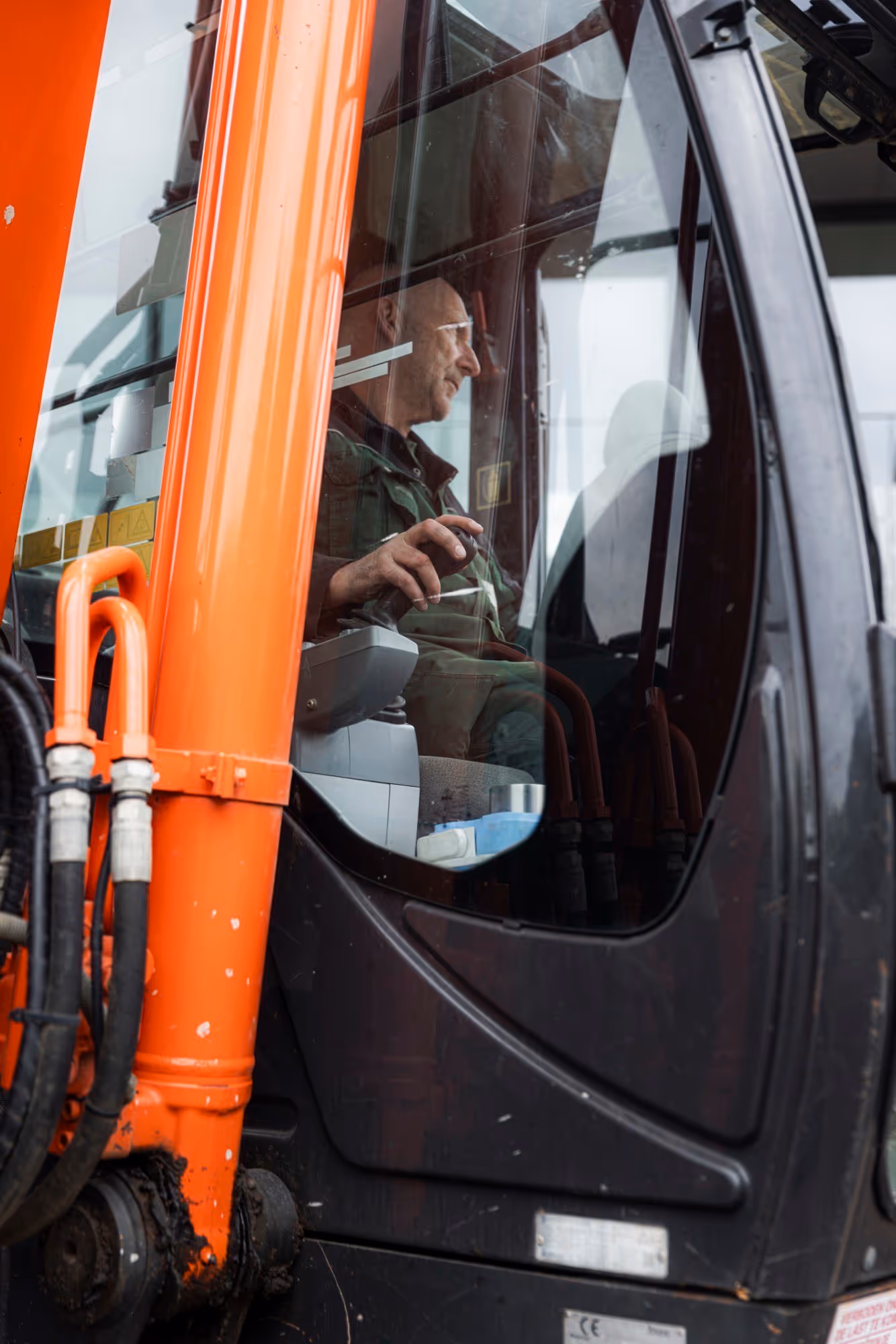 Man bestuurt een graafmachine, zichtbaar door het raam van de cabine, met een oranje mechanisch onderdeel op de voorgrond.