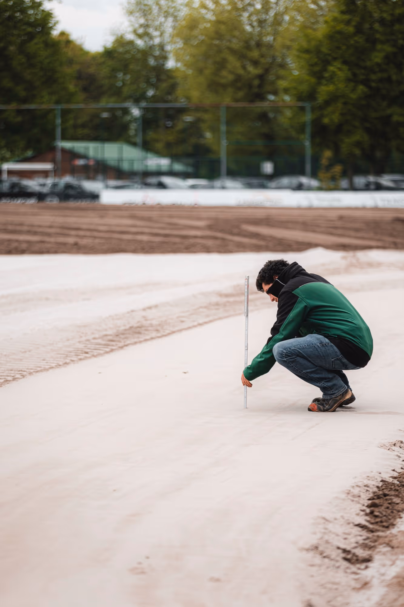 Man in een groene jas hurkt en meet met een meetlat op een zand- of bouwterrein.