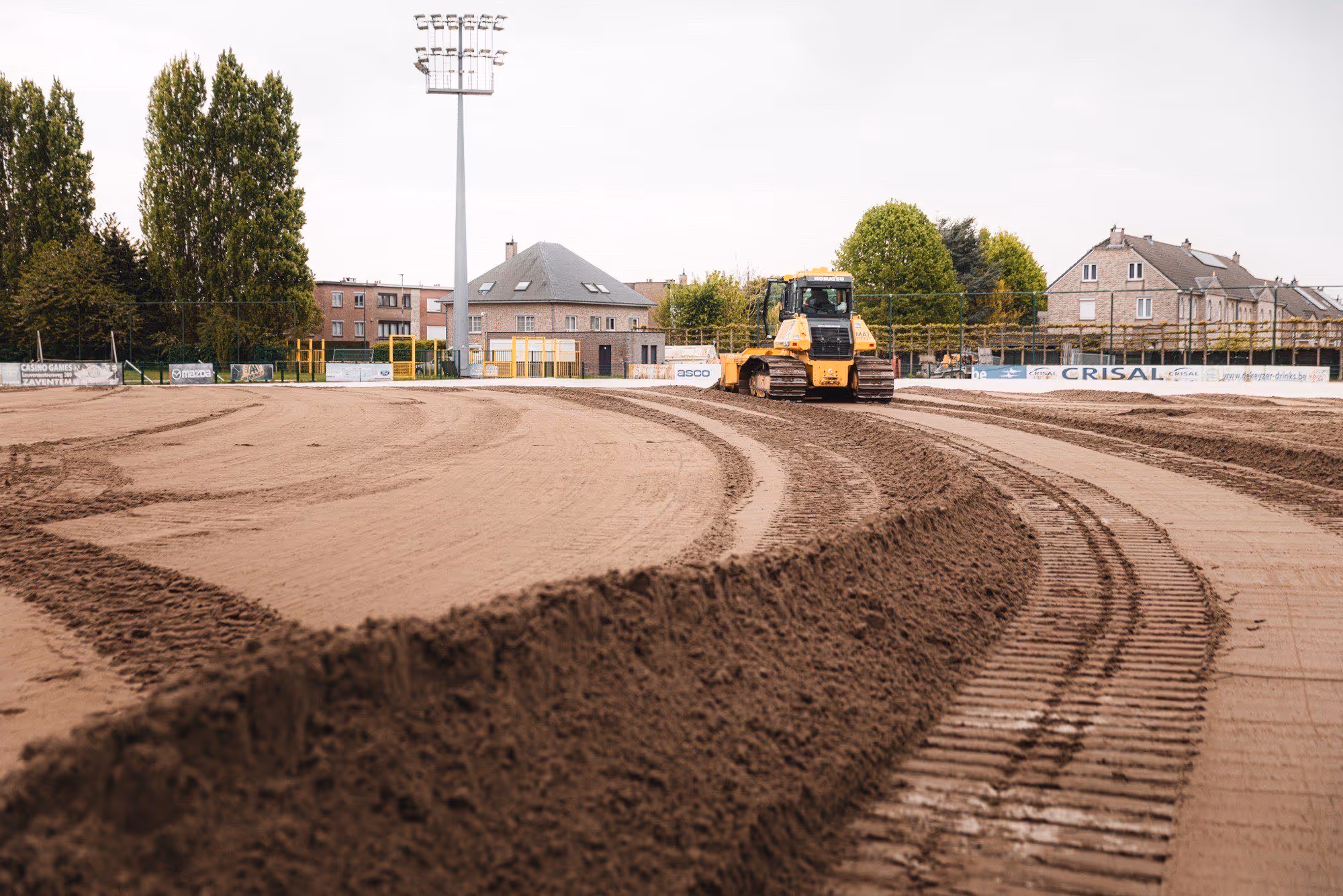 Gele bulldozer maakt een gebogen baan op een zandveld, omringd door huizen, bomen en een hoge lichtmast.
