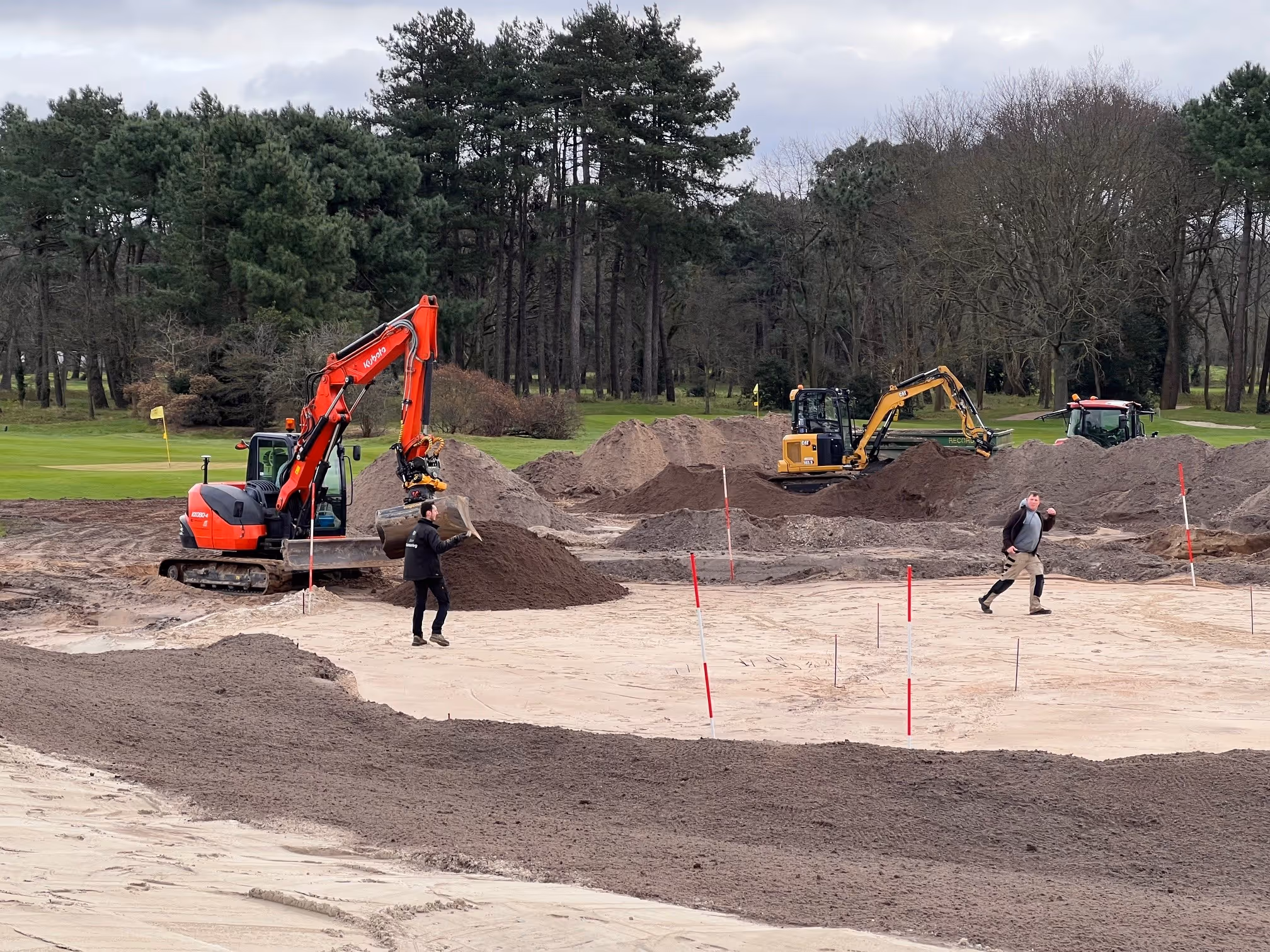 Twee graafmachines en twee arbeiders aan het werk op een zandige bouwplaats nabij een golfbaan met bomen op de achtergrond.