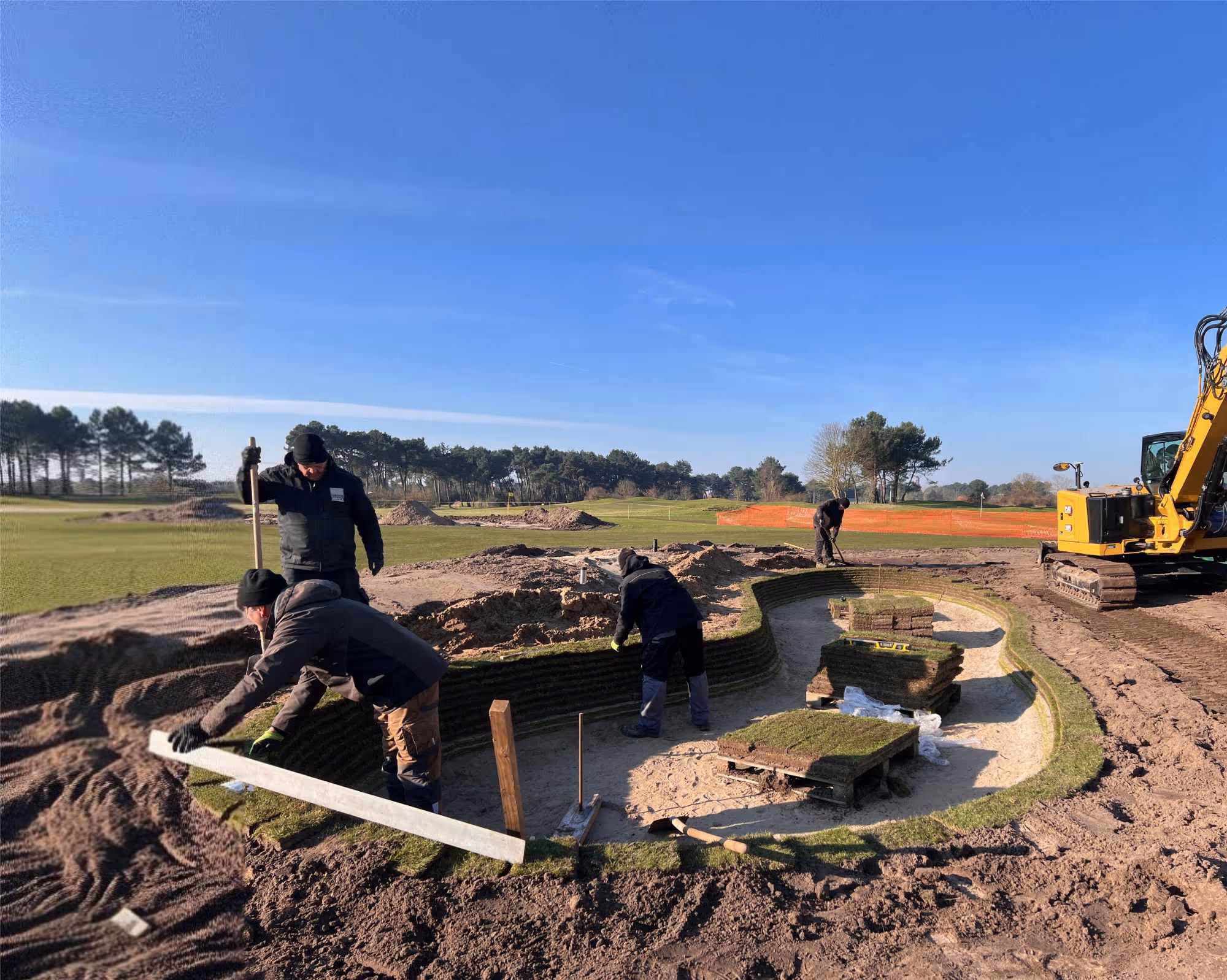 Vier arbeiders leggen grasmatten in de vorm van een bunker op een golfbaan onder een blauwe heldere hemel.