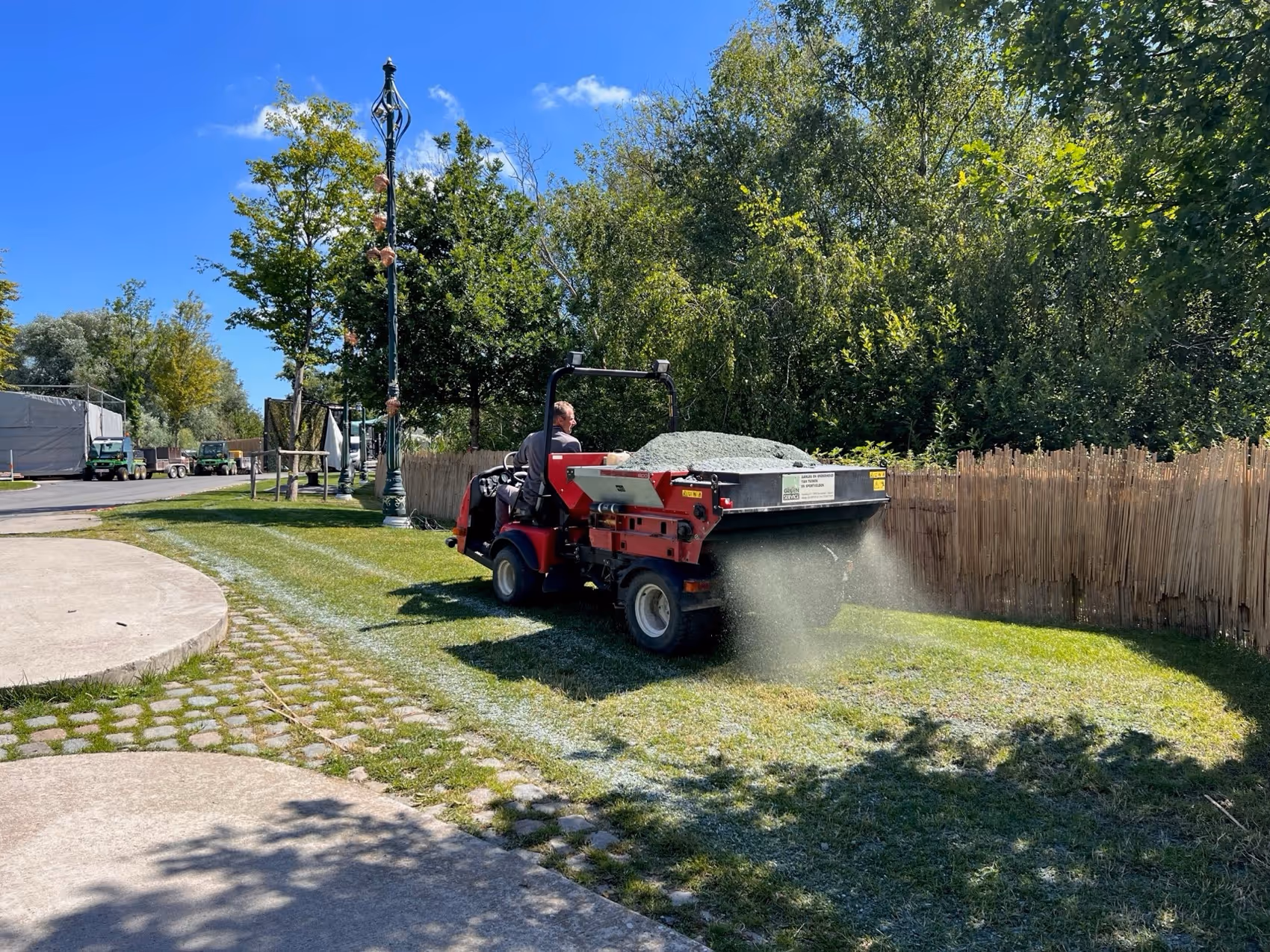 Man op een voertuig strooit kalk op een grasveld langs een pad onder een heldere blauwe lucht.
