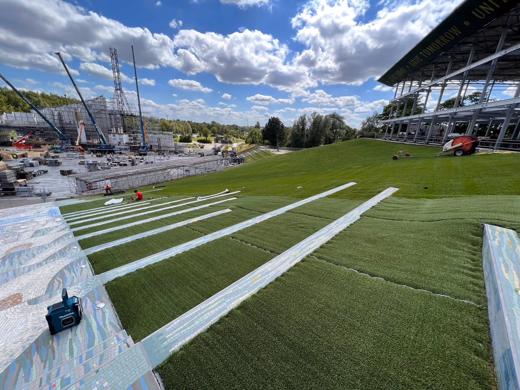 Mensen installeren kunstgras op een helling met een bouwplaats en kraan op de achtergrond onder een partly bewolkte blauwe lucht.