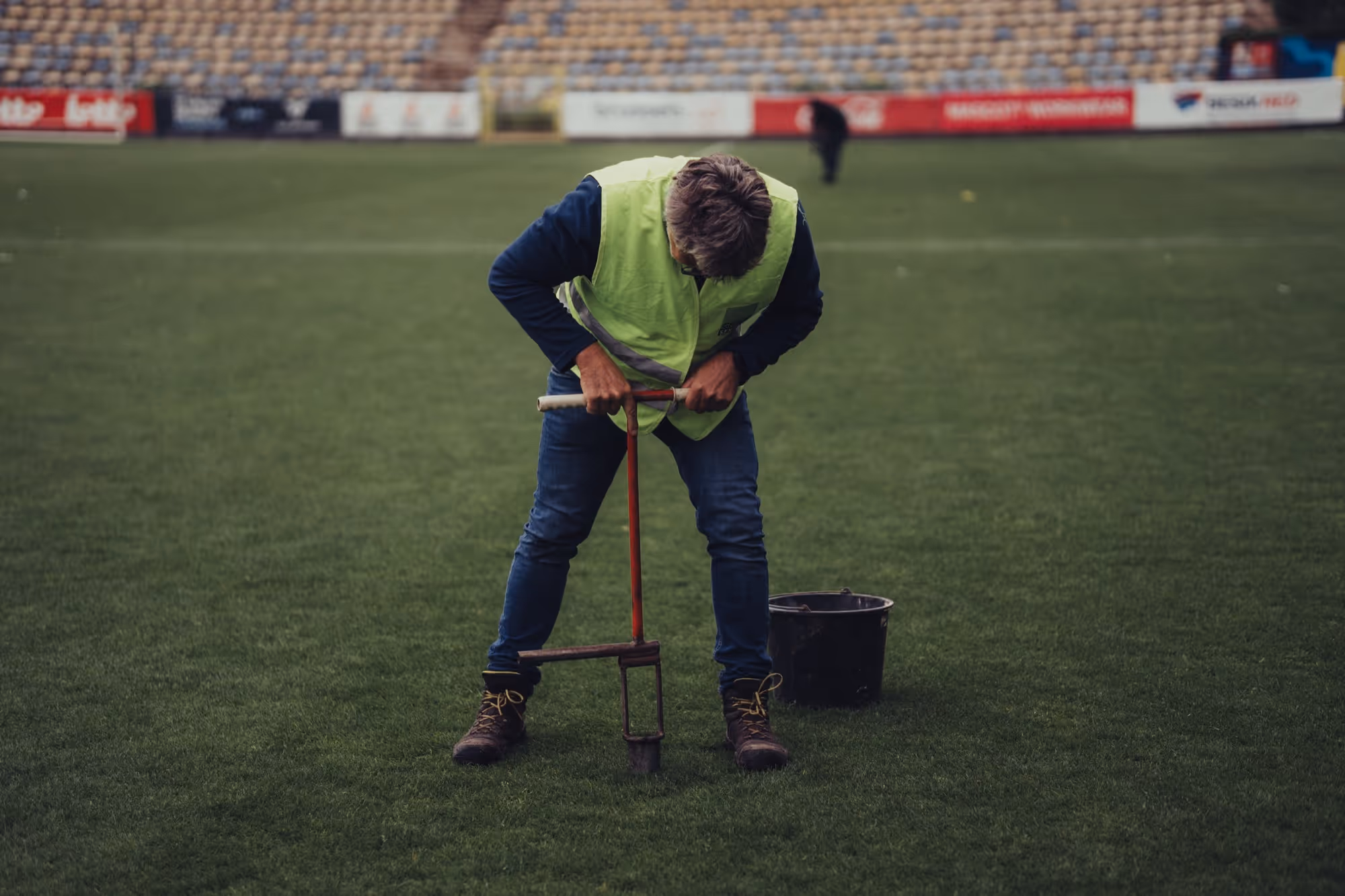 Man in een fluorescerend hesje die met een grondboor in een grasveld werkt, met een emmer naast hem.