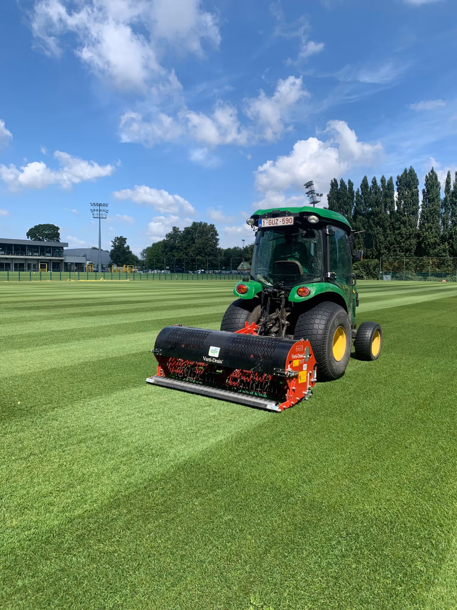 Groene tractor met een beluchtingsmachine onderhoudt een strak gemaaid sportveld onder een blauwe lucht.