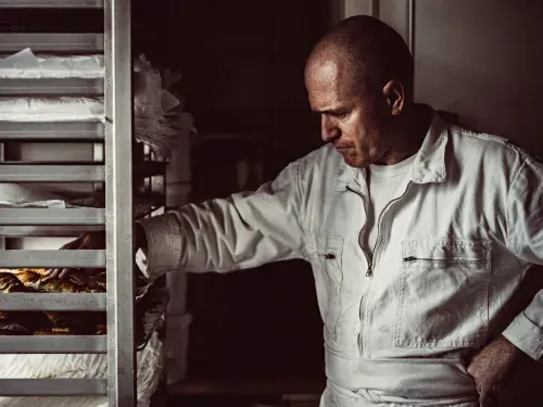 Christophe Vasseur en tenue de boulanger observant des viennoiseries sur une étagère de fournil, concentré sur le contrôle de la cuisson.