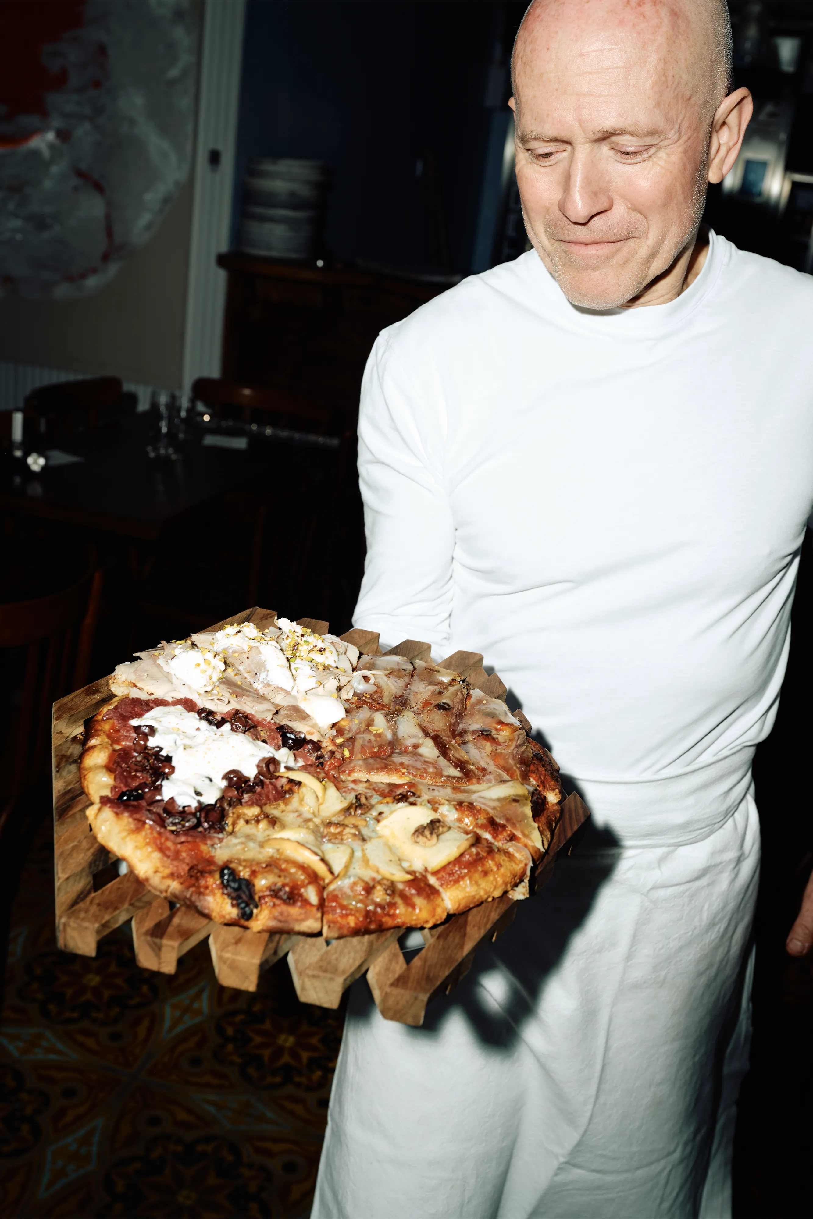 Christophe Vasseur en tenue blanche tient une grande pizza aux garnitures variées sur un plateau en bois.