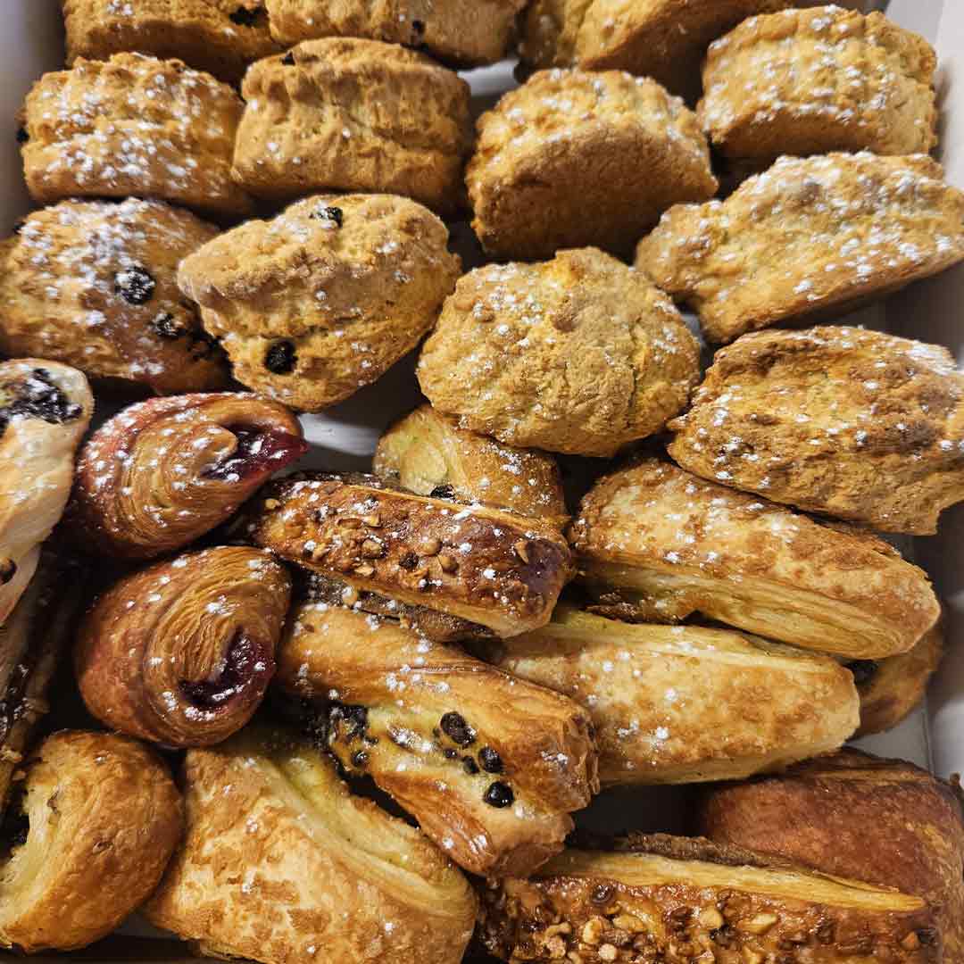 Assorted golden-brown pastries with powdered sugar and raisins, including scones and rolls, arranged closely together.