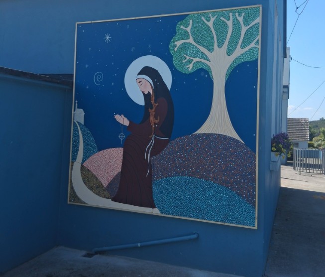 Mural of a woman in a dark robe praying with a rosary under a halo by a tree on curved hills.