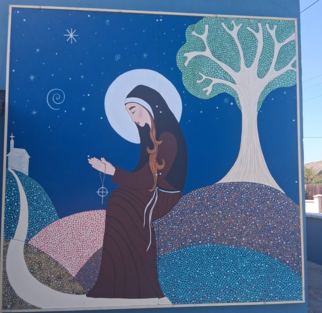 Mural of a woman in a dark robe holding rosary beads against a starry night background with hills and a tree.