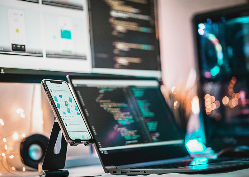 Smartphone on stand displaying flowchart diagrams next to laptop and monitor showing colorful code in a dimly lit workspace.