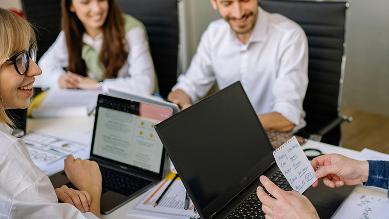 Three people in a meeting around a table with laptops and notes, one person holding a card with handwritten tasks.