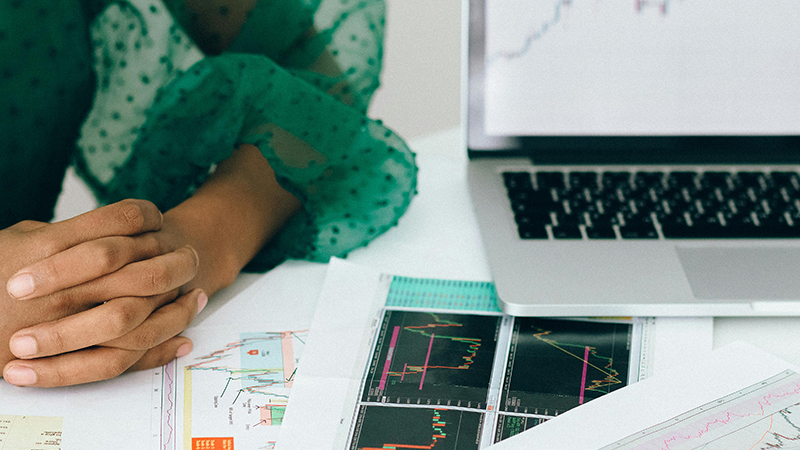 Hands resting on a desk with printed financial charts beside a laptop displaying a stock chart.