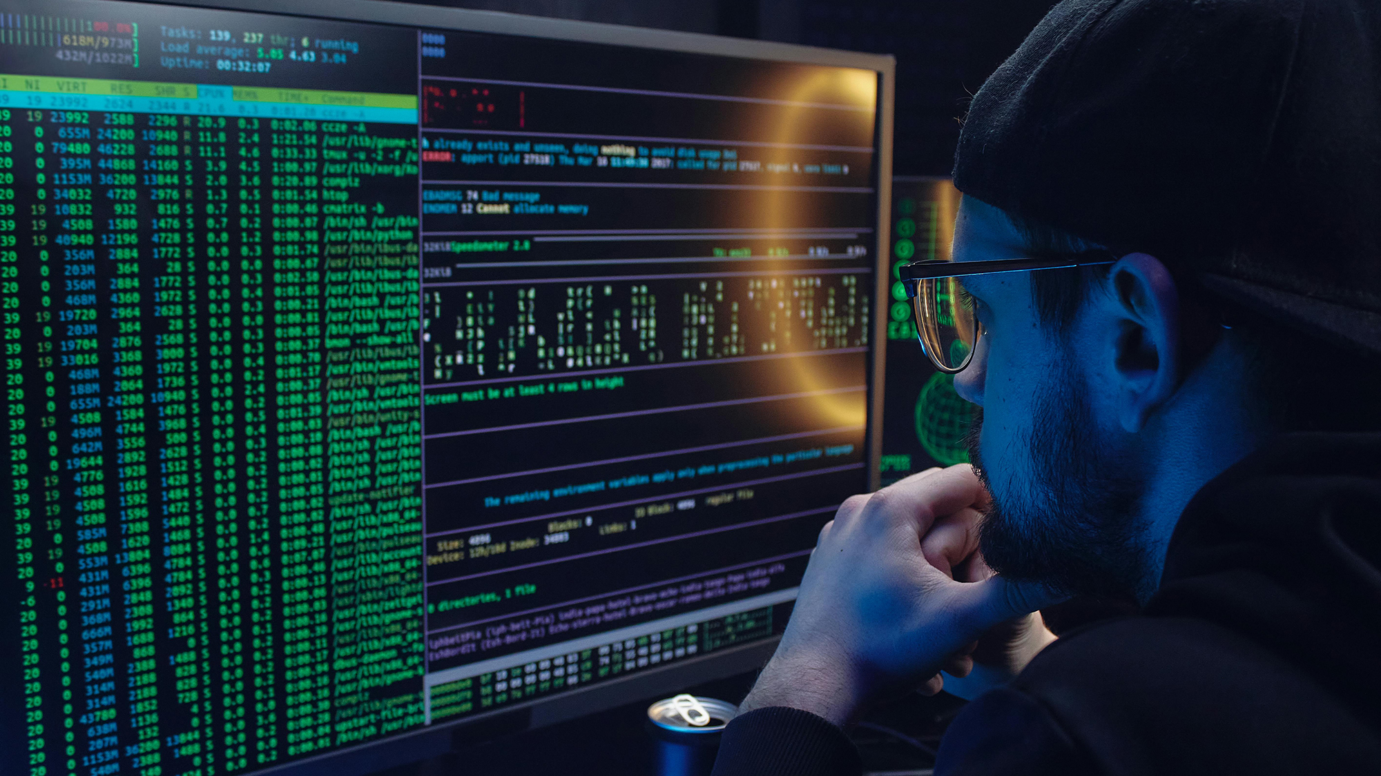 Man wearing glasses and a cap attentively viewing computer screen filled with green and white terminal code lines.