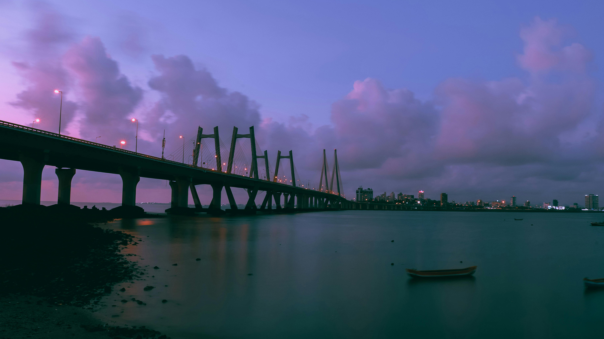 Wide view of a long cable-stayed bridge over calm water at dusk with city lights and purple clouds in the background.