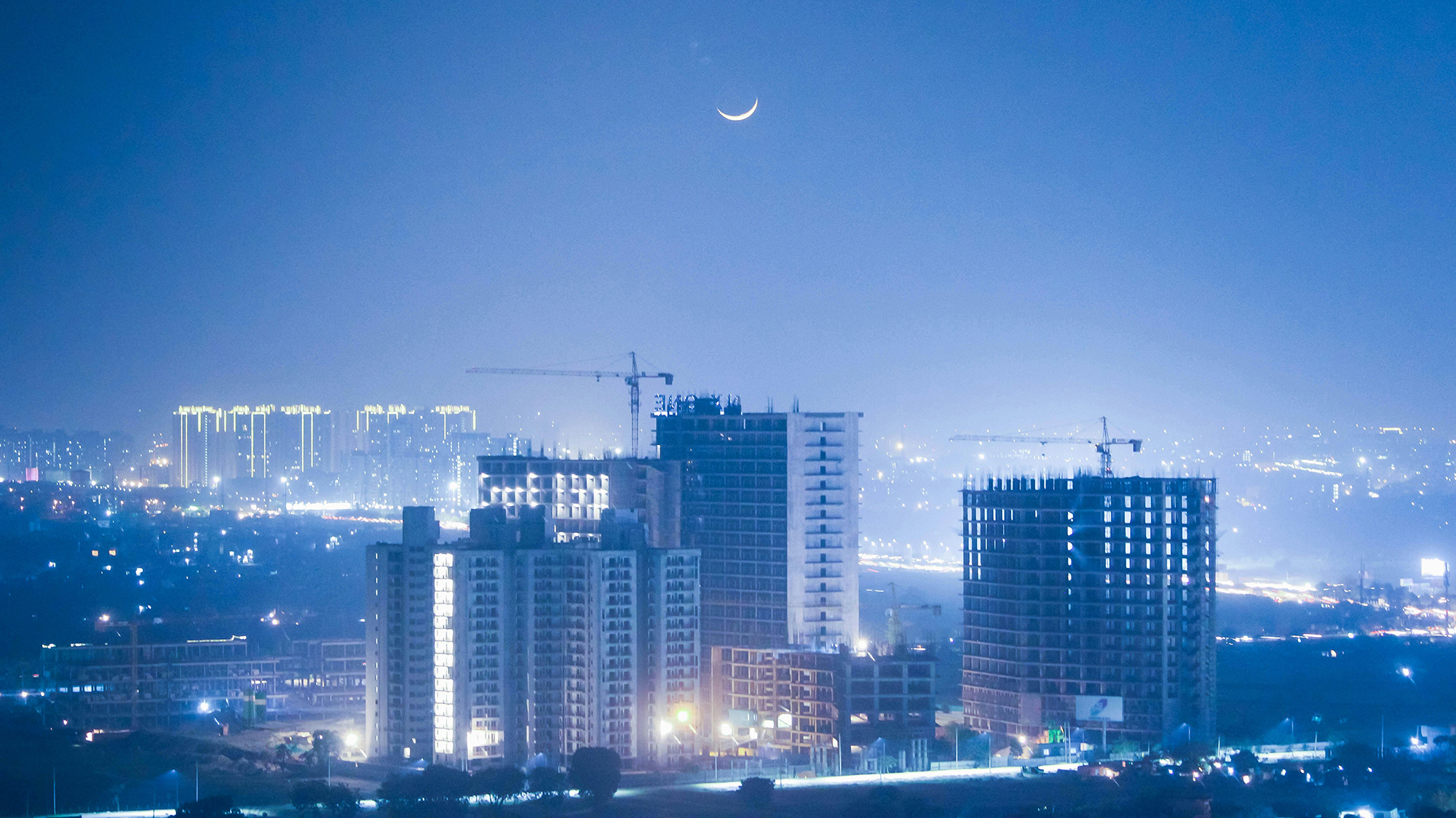 Cityscape at night with illuminated high-rise buildings under construction and a crescent moon in a clear blue sky.