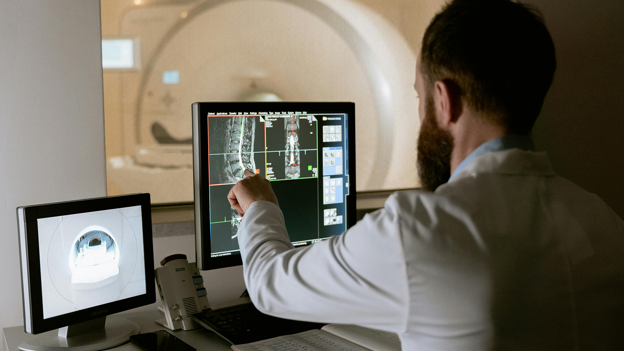 Doctor pointing at spinal MRI images on a computer screen next to an MRI machine control monitor.