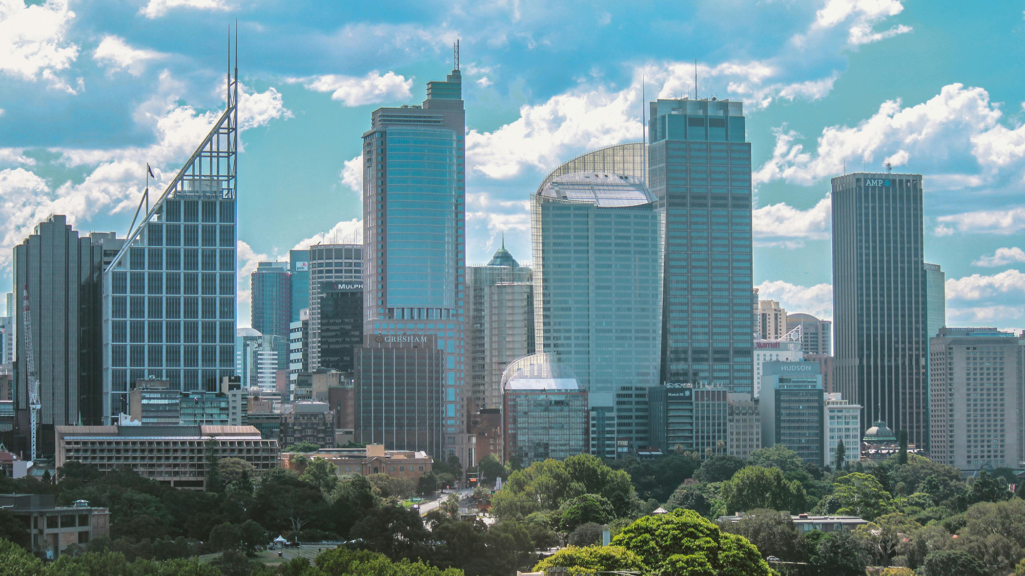 City skyline with modern high-rise buildings under a blue sky with scattered clouds, with a green park in the foreground.