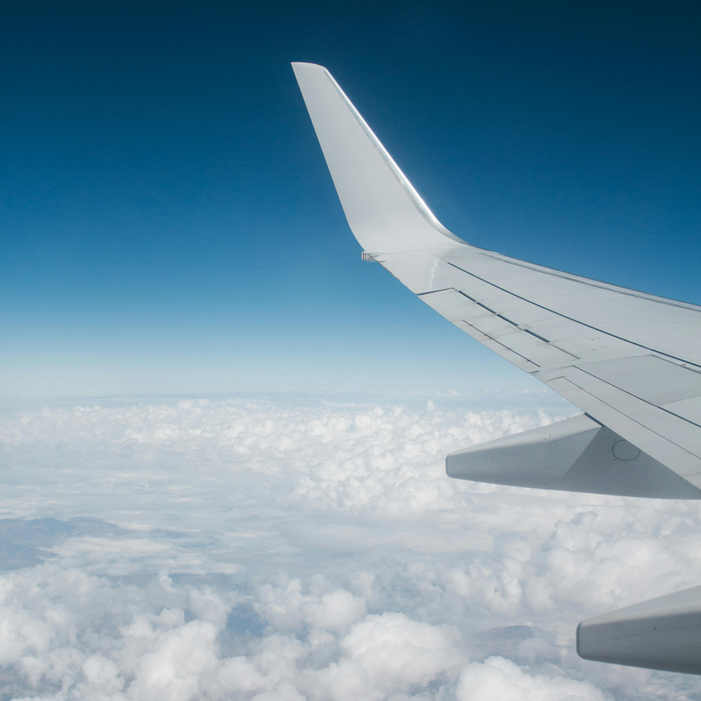 View of an airplane wing flying above fluffy white clouds under a clear blue sky.