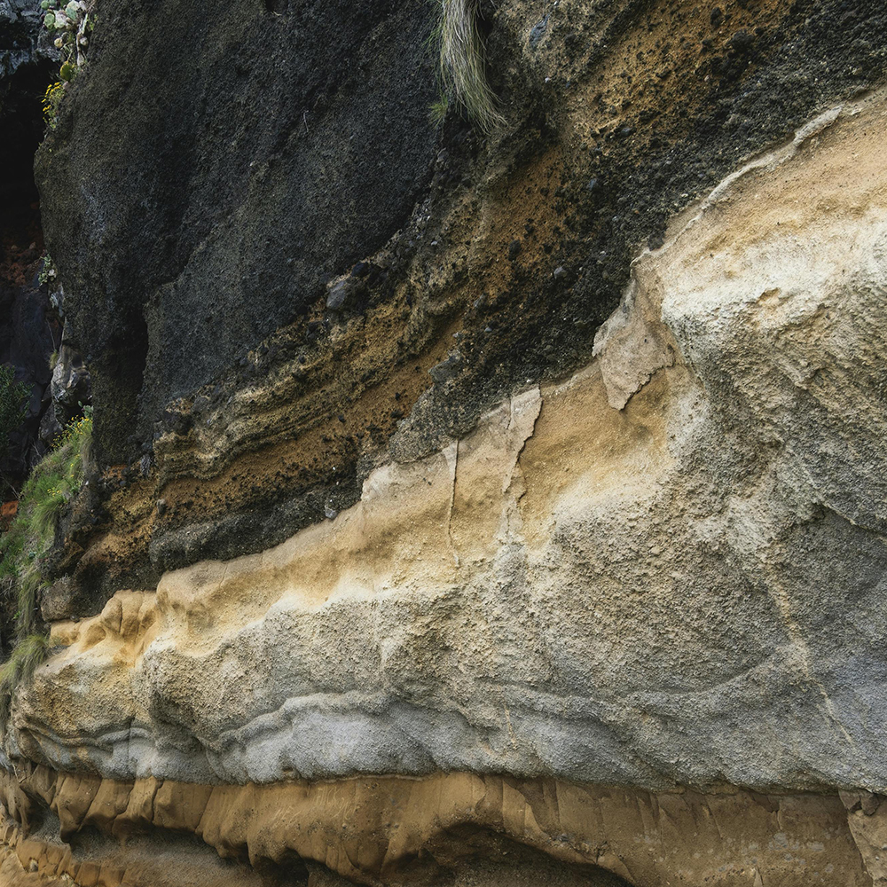 Close-up of a multi-layered rock formation showing distinct textured strata in shades of black, brown, beige, and gray.