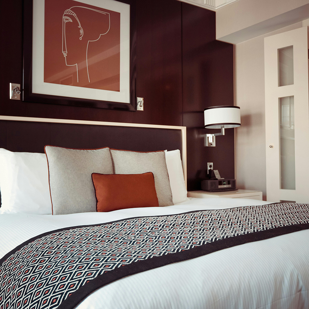 Neatly made bed with white linens, three decorative pillows in beige and brown, and a patterned runner, against a dark wood paneled wall with abstract face art and a wall-mounted lamp.