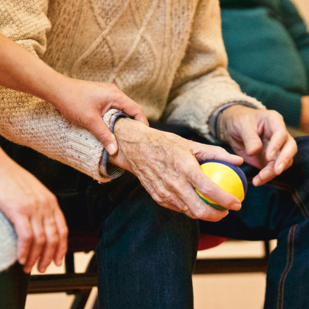 Younger hand gently holding older person's arm who is holding a yellow and blue ball.