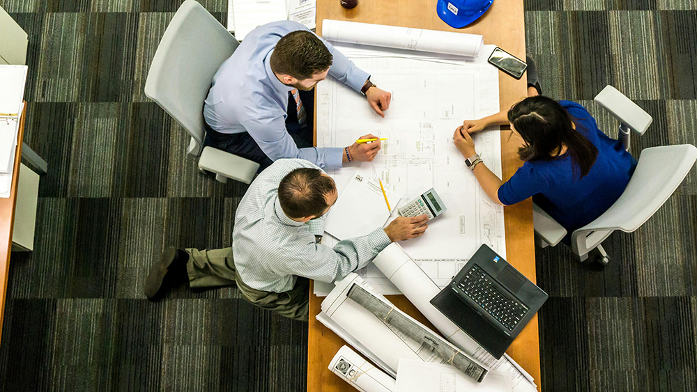 Three professionals reviewing architectural blueprints at a table with a laptop, calculator, and rolled plans.