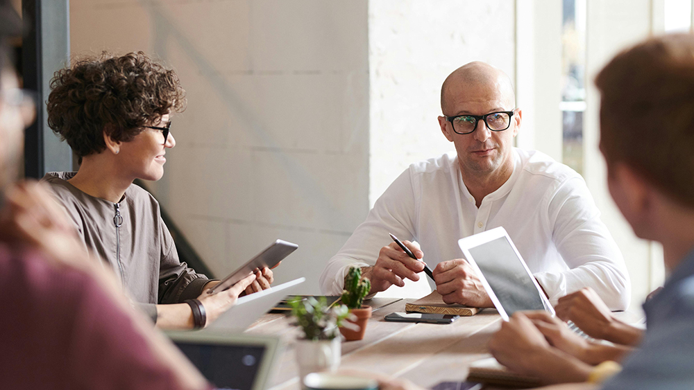 Four people in a meeting around a table with tablets and laptops, engaged in discussion.