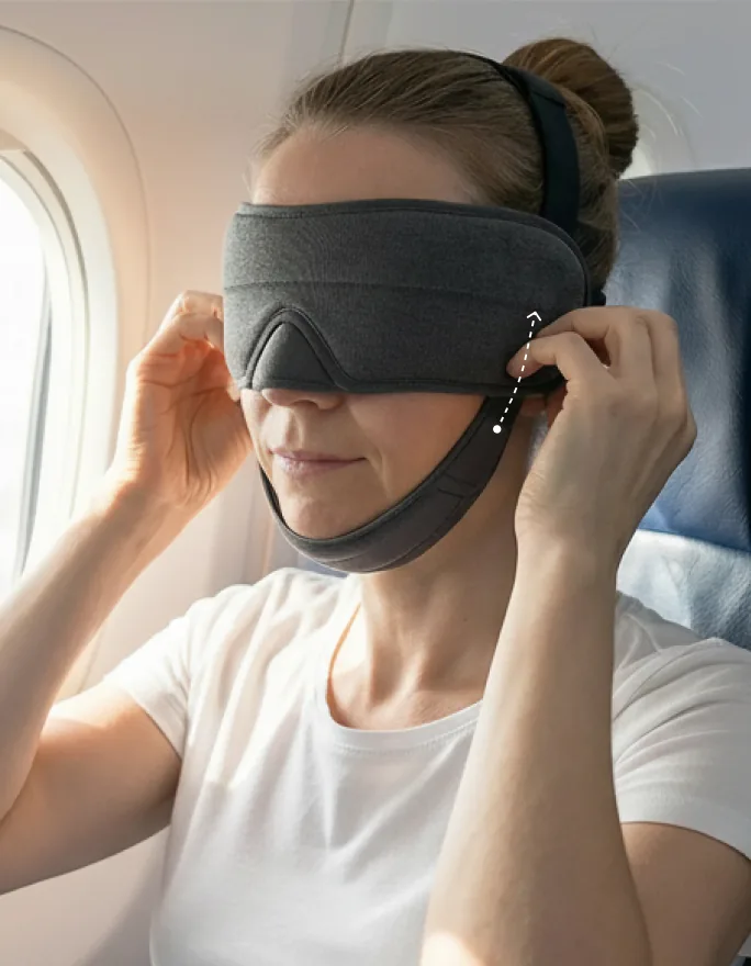 Woman on a flight adjusting the Sleepmaxxer sleep mask's strap for a secure, custom fit while traveling