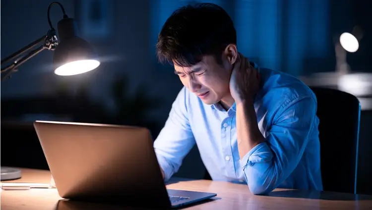 A man working at a desk late at night, gripping the back of his neck in physical discomfort from severe neck stiffness.