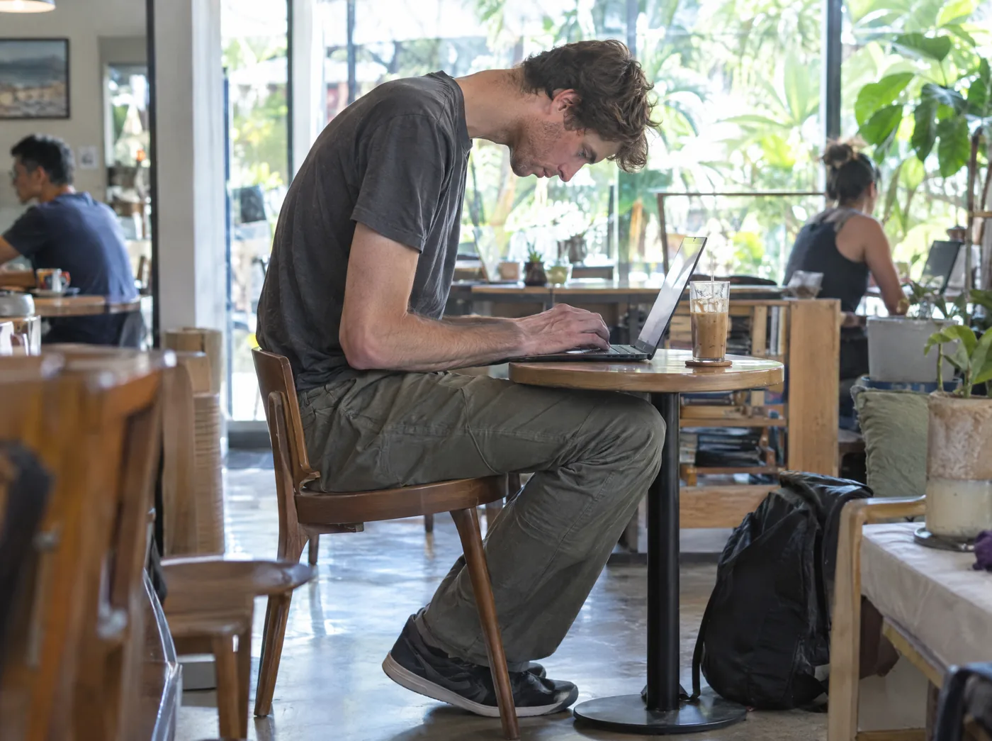 Side view of a tall man hunched over a small laptop at a low cafe table, his head well in front of his shoulders