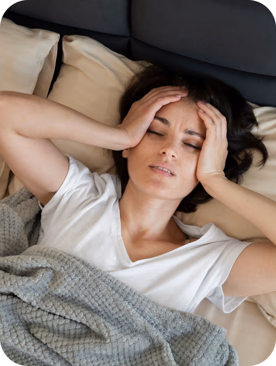 Frau liegt im Bett mit geschlossenen Augen und hält sich schmerzhaft den Kopf.
