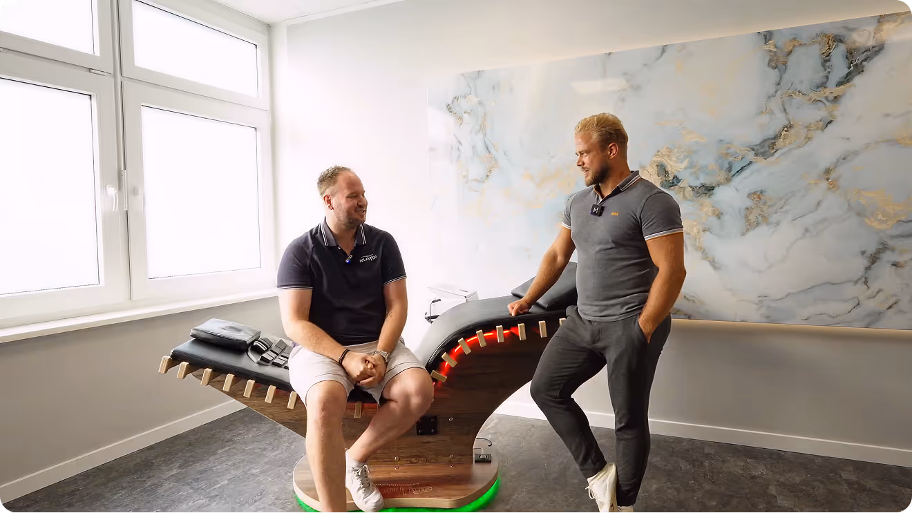 Zwei Männer unterhalten sich in einem hellen Raum mit großen Fenstern, einer sitzt auf einem modernen Entspannungsstuhl mit beleuchteter Basis, der andere lehnt daneben.