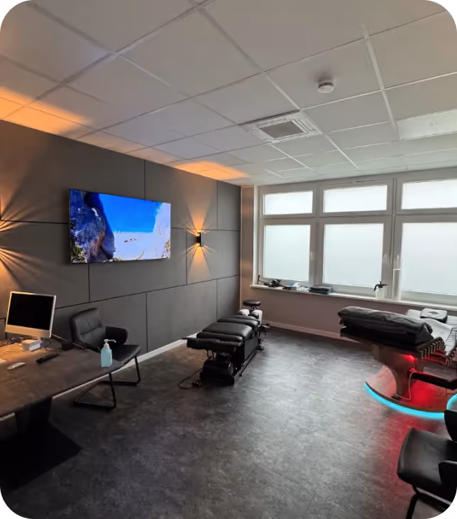 Moderne Behandlungskabine mit grauer Wand, Deckenbeleuchtung, Fernseher, Schreibtisch mit Computer und Handdesinfektionsmittel, Behandlungsbett und großer Fensterfront mit Tageslicht.