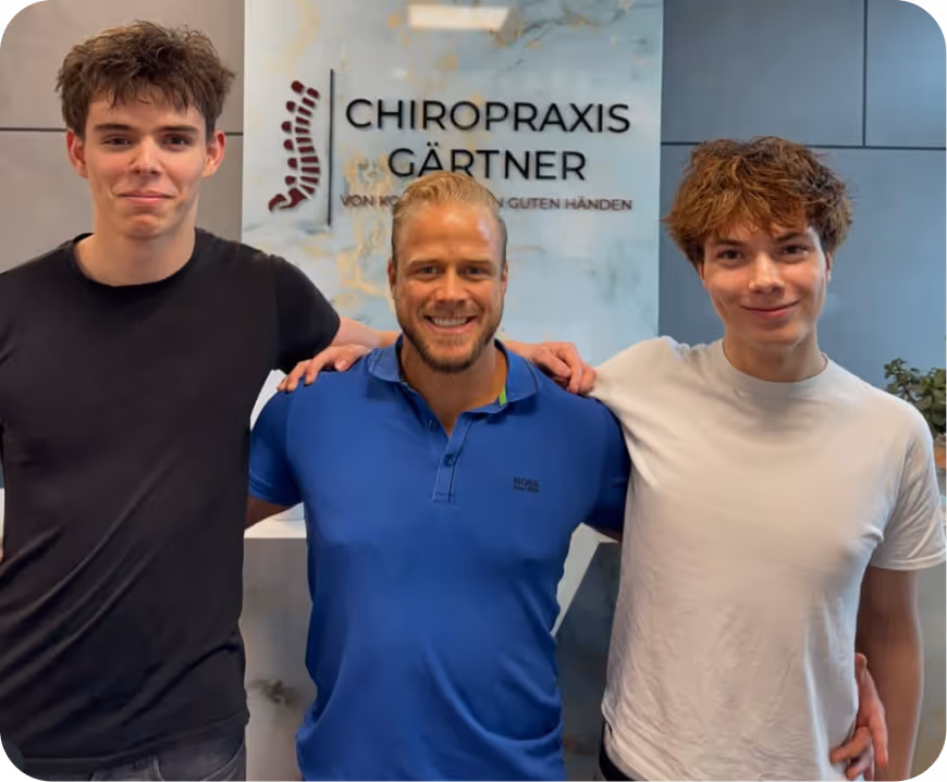 Drei Männer stehen lächelnd vor einer Wand mit Schriftzug 'Chiropraxis Gärtner', der Mann in der Mitte trägt ein blaues Poloshirt.
