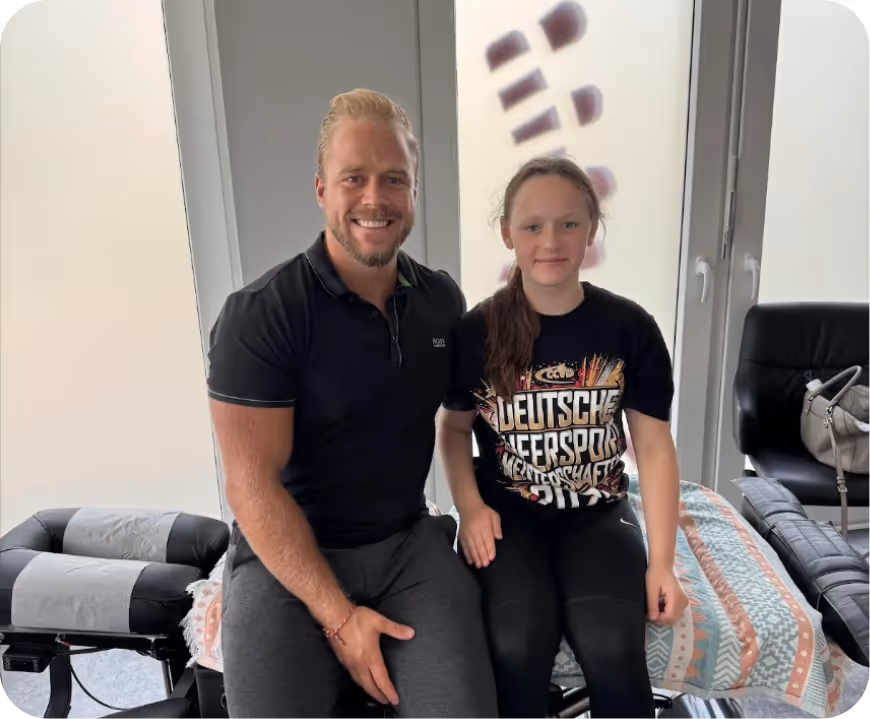 Zwei lächelnde Personen, ein Chiropraktor und ein Mädchen, sitzen nebeneinander auf einer Behandlungsliege vor einem Fenster mit weißen Vorhängen.