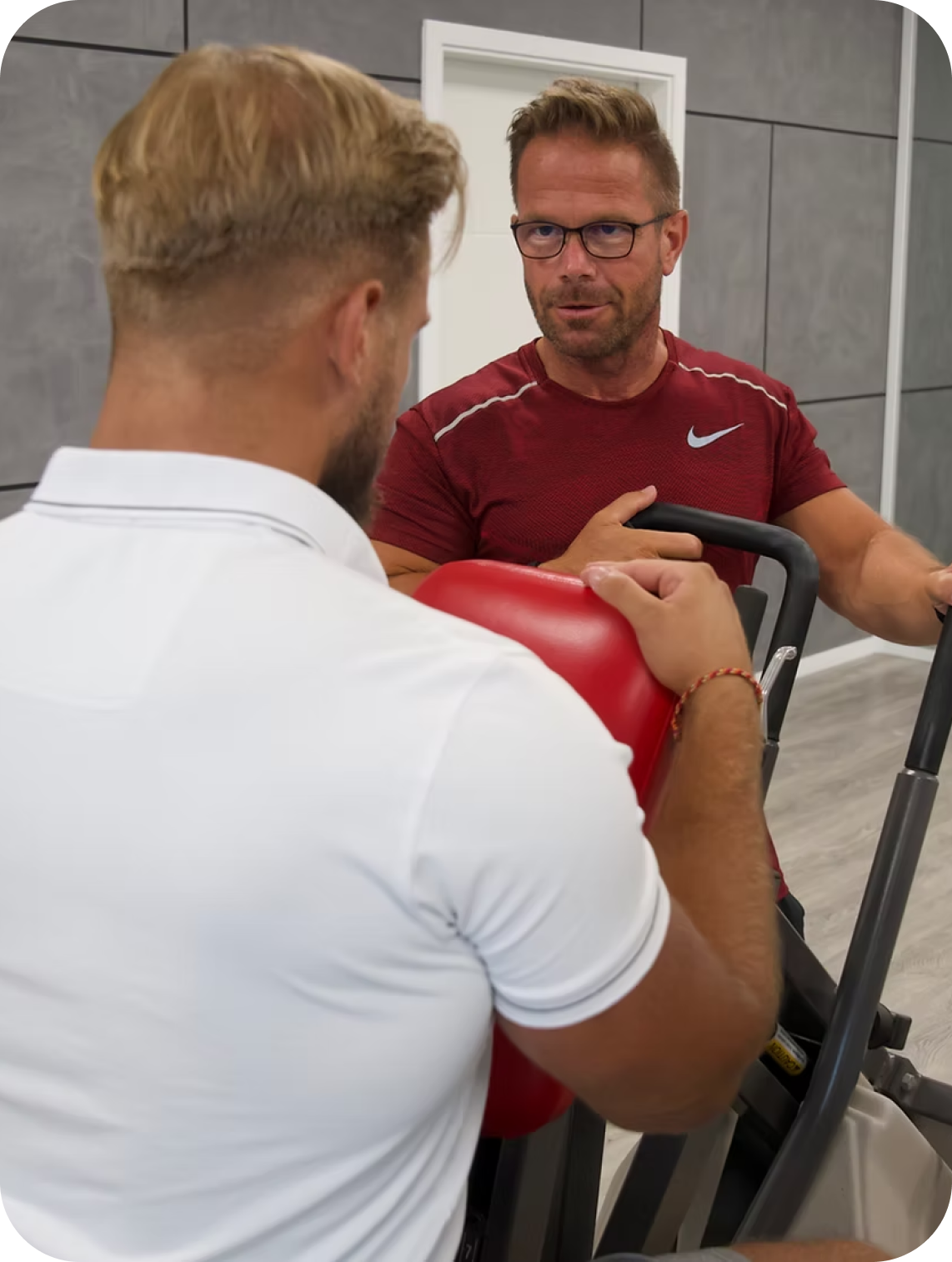 Ein Fitnesscoach in rotem Shirt erklärt einem Mann in weißem Poloshirt die Benutzung eines Trainingsgeräts mit rotem Polster.