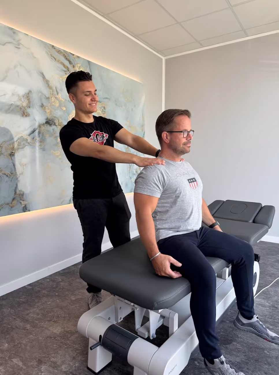 Ein Mann sitzt auf einer Behandlungsliege, während ein anderer Manual Therapeut ihm die Schultern massiert in einem modernen Behandlungsraum.